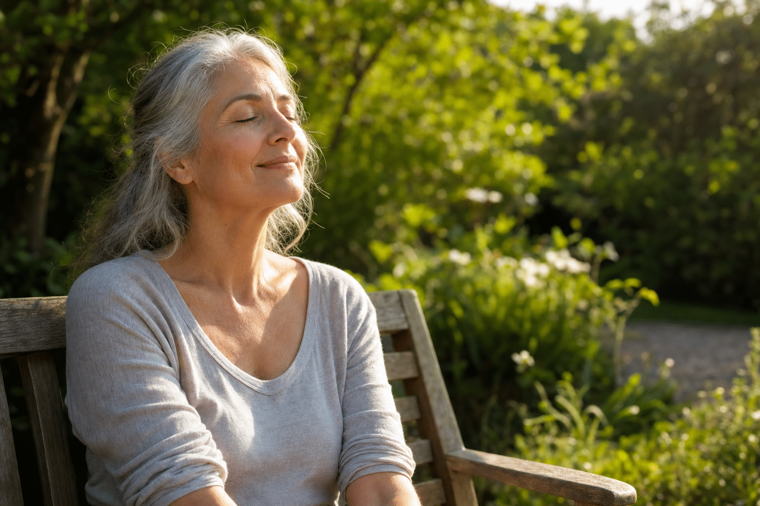 A woman in her late fifties with silver hair sitting outdoors in gentle morning sunlight with her forearms bare, eyes closed and relaxed, in a green garden setting